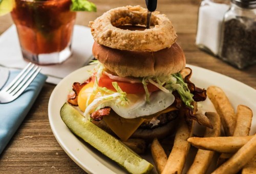 A hearty burger topped with bacon, cheese, lettuce, tomato, and an onion ring, served with fries and a pickle.