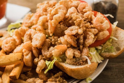 A close-up of a shrimp sandwich piled high with fried shrimp, lettuce, and tomato, served with fries on the side.