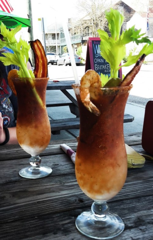 Two tall glasses of Bloody Marys garnished with celery, shrimp, and crispy bacon, sitting on a wooden table outdoors.