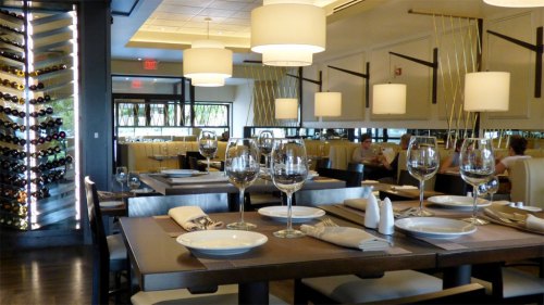 Elegant restaurant interior with tables set for dining, wine display, and soft lighting. Guests are seated in the background.