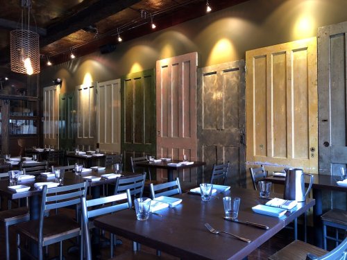 A cozy restaurant interior featuring wooden tables, chairs, and a wall adorned with colorful vintage doors.