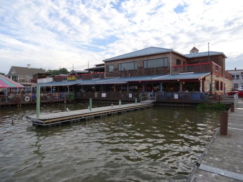 A waterfront restaurant with a wooden deck, surrounded by water and a cloudy sky. Colorful awnings are visible.