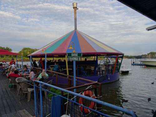 Colorful barge restaurant on a lake, with outdoor seating and people enjoying the view under a cloudy sky.