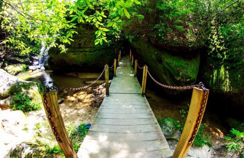 A wooden bridge with chains crosses a serene, narrow creek surrounded by lush greenery and rocky cliffs.