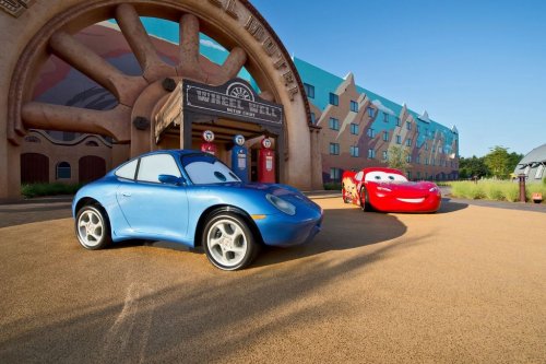 Two animated cars, a blue sports car and a red race car, parked in front of a whimsical building in a sunny setting.