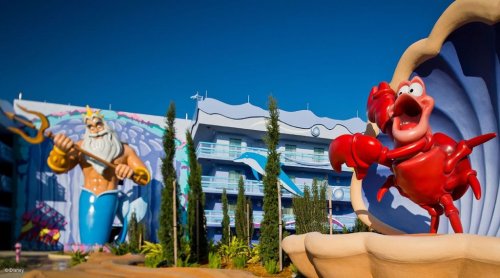 A vibrant scene featuring a large statue of King Triton and Sebastian the crab outside a colorful hotel.