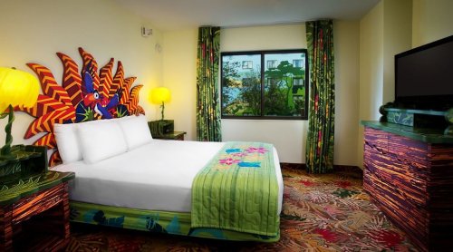 Brightly decorated hotel room with a large bed, tropical-themed decor, and colorful furnishings. Large window with greenery outside.