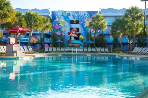 A vibrant pool area with palm trees and a mural of a mermaid, surrounded by lounge chairs and colorful decor.