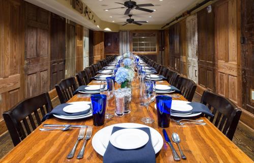 A long wooden dining table set with plates, glasses, and blue napkins, surrounded by wooden chairs in a rustic room.