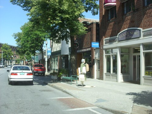 A street view of a small town with shops, a statue, and parked cars under clear blue skies.