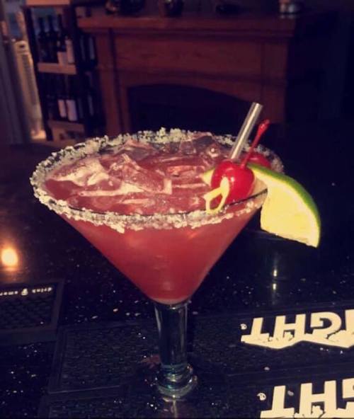 A vibrant cocktail in a glass with a salted rim, garnished with a cherry and lime wedge, on a dark bar surface.
