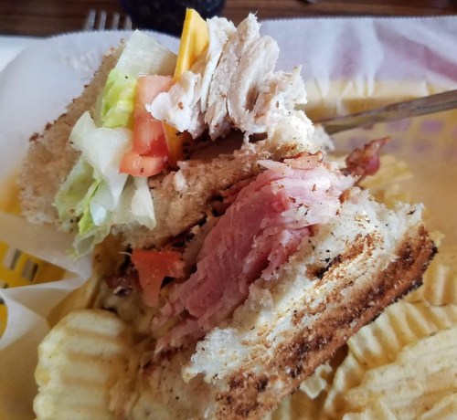 A close-up of a layered sandwich with turkey, ham, lettuce, tomato, and cheese on toasted bread, served with ridged chips.