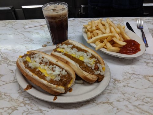 A plate with two chili cheese hot dogs topped with onions, served with fries and a glass of soda on a marble table.