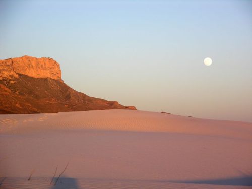 A serene desert landscape at sunset, featuring a mountain and a full moon in the sky above soft, sandy dunes.