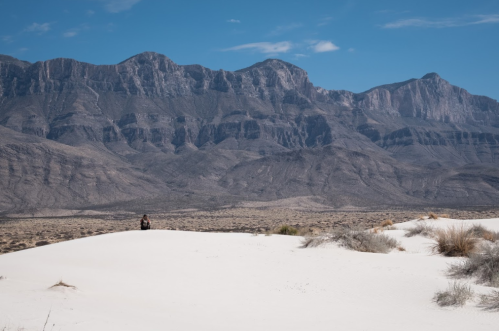 A lone figure sits on a white sand dune with rugged mountains in the background under a clear blue sky.