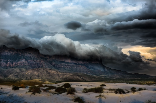Dramatic clouds loom over rugged mountains, casting shadows on a sandy landscape under a moody sky.
