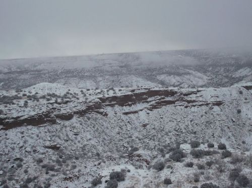 A snowy landscape with rocky cliffs and sparse vegetation under a cloudy sky.