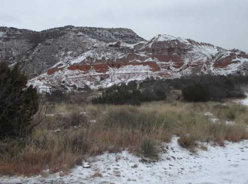 Snow-dusted red rock formations rise above a grassy landscape under a cloudy sky.