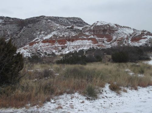 Snow-dusted red rock formations rise above a grassy landscape under a cloudy sky.