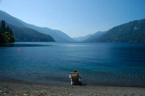 A person sits in a chair by a calm lake, surrounded by mountains under a clear blue sky.