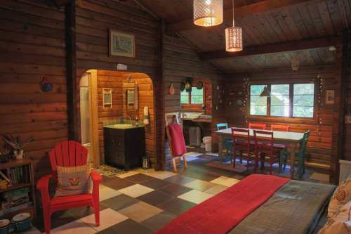 Cozy wooden cabin interior featuring a living area, dining table, and kitchenette with colorful furniture and warm lighting.