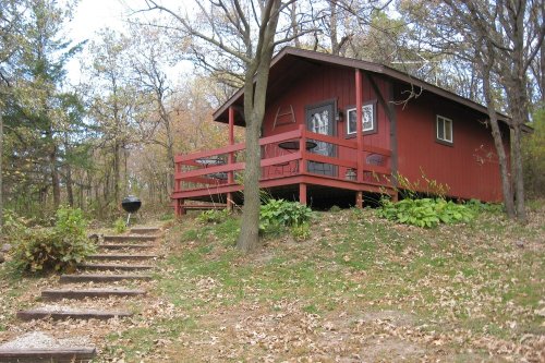 A red cabin with a porch surrounded by trees and autumn foliage, with stone steps leading up to it.