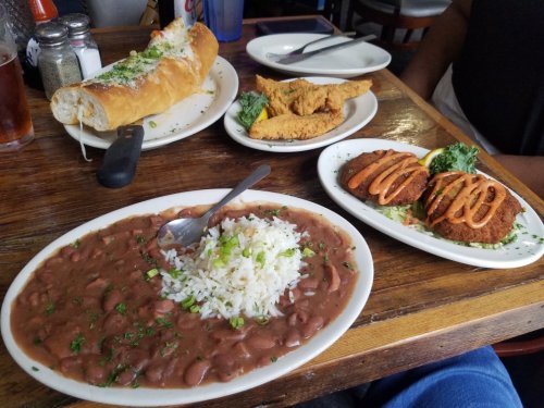 A table with a plate of rice and beans, fried fish, a sandwich, and two patties drizzled with sauce.