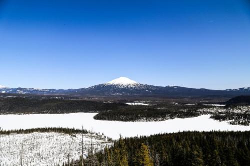 Snow-covered mountain rises above a frozen lake, surrounded by dense forests under a clear blue sky.