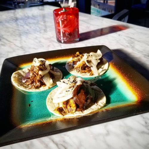 Three tacos on a colorful plate, topped with meat, onions, and sauce, with a red drink in the background.
