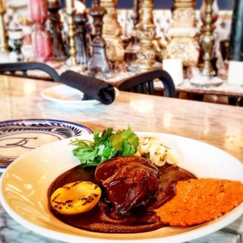 A beautifully plated dish featuring meat, grilled peach, herbs, and a crispy tortilla, set against a stylish restaurant backdrop.