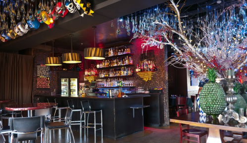A vibrant bar interior with colorful masks, a well-stocked shelf, and decorative lighting, creating a lively atmosphere.