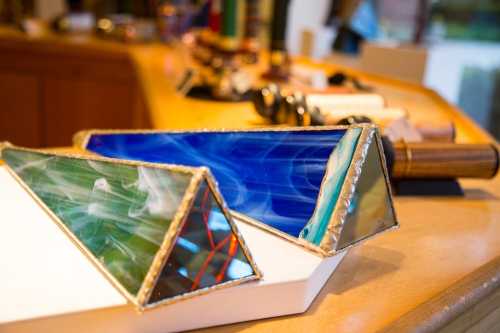 Colorful stained glass pyramids in green and blue, displayed on a wooden surface with various tools in the background.