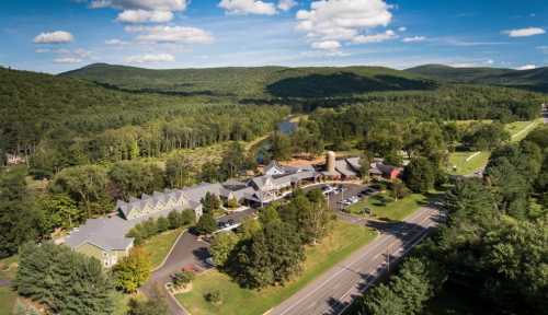 Aerial view of a hotel surrounded by lush green mountains and a river, with a clear blue sky above.