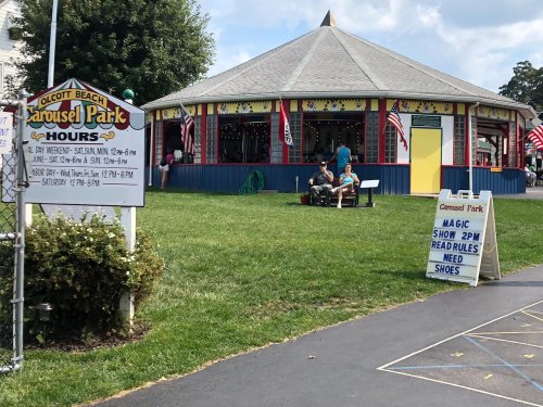 A colorful carousel building at Olcott Beach, with a sign listing hours and people enjoying the park on a sunny day.