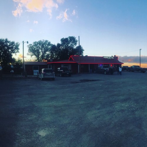 A rustic building with red neon lights at dusk, surrounded by parked cars and trees under a colorful sky.