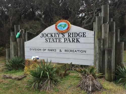 Sign for Jockey's Ridge State Park, featuring the park's name and logo, surrounded by greenery and wooden posts.