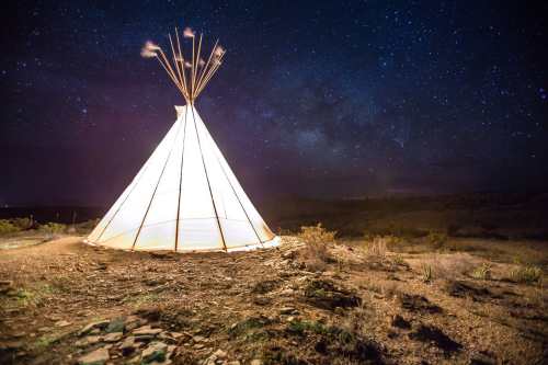 A lit teepee under a starry night sky, surrounded by desert landscape.
