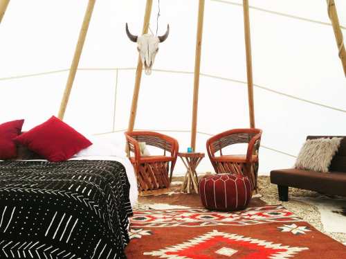Cozy interior of a teepee with a bed, two chairs, a small table, and decorative rugs. A skull hangs from the ceiling.