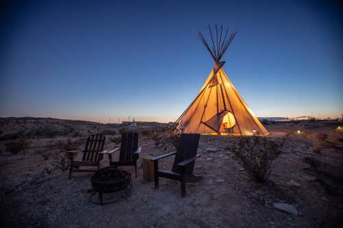 A lit teepee at dusk, surrounded by wooden chairs and a fire pit in a desert landscape.