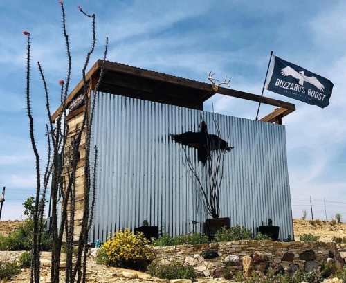A rustic building with a metal exterior, featuring a flag, antlers, and cacti in a desert landscape.