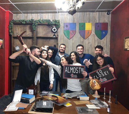 A group of eight friends celebrating in an escape room, holding signs that say "ALMOST!!!" and "SO CLOSE!"