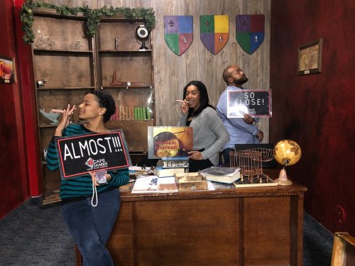 Three people in an escape room holding signs that say "ALMOST!!!" and "SO CLOSE!" with a decorated background.
