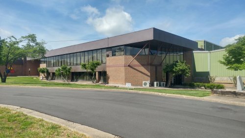 A modern brick building with large windows, surrounded by trees, located on a curved road under a blue sky.