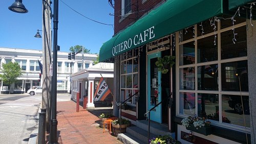 A sunny street view of Quiero Cafe with an open sign, planters, and nearby buildings in a charming town setting.