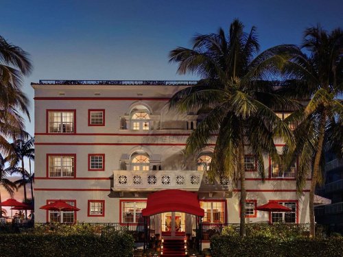A charming hotel with red accents, palm trees, and a welcoming entrance, illuminated at dusk.