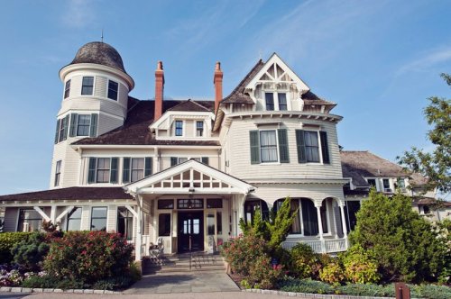 A large, elegant Victorian-style house with a turret, surrounded by lush greenery and colorful flowers under a clear blue sky.