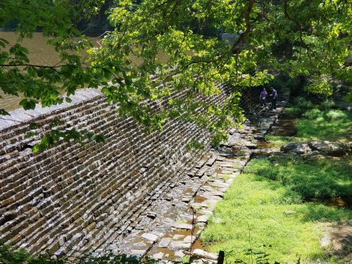 A stone wall along a riverbank, with greenery and two people walking nearby under a sunny sky.