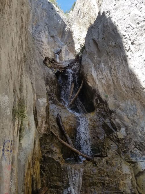 A narrow rocky canyon with a waterfall cascading down, surrounded by steep cliffs and scattered logs.