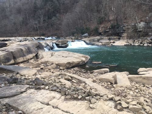 A serene river scene with flowing water over rocks, surrounded by trees and a rocky shoreline.