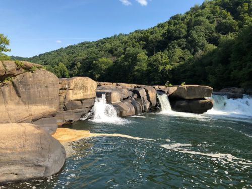 A serene river scene with cascading waterfalls surrounded by lush green hills and rocky formations under a clear blue sky.